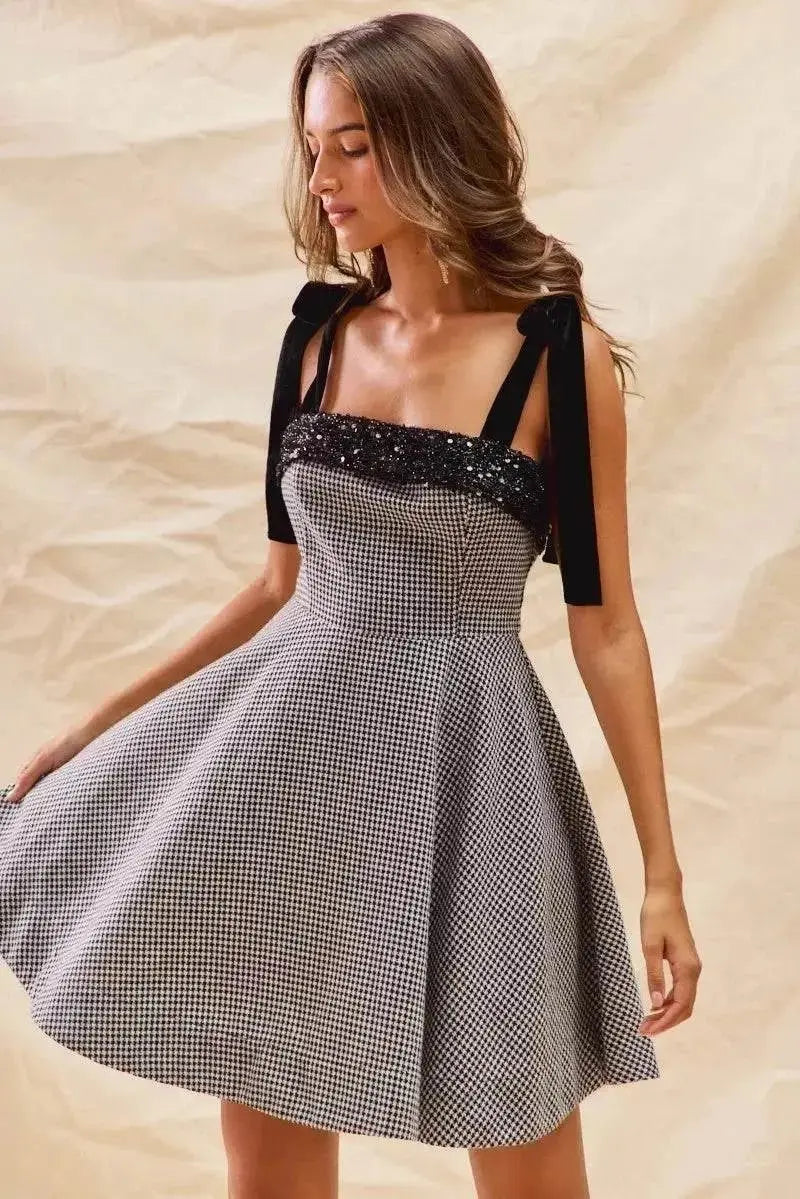 Woman wearing a houndstooth fit flare mini dress with shoulder tie straps and black sequin detail