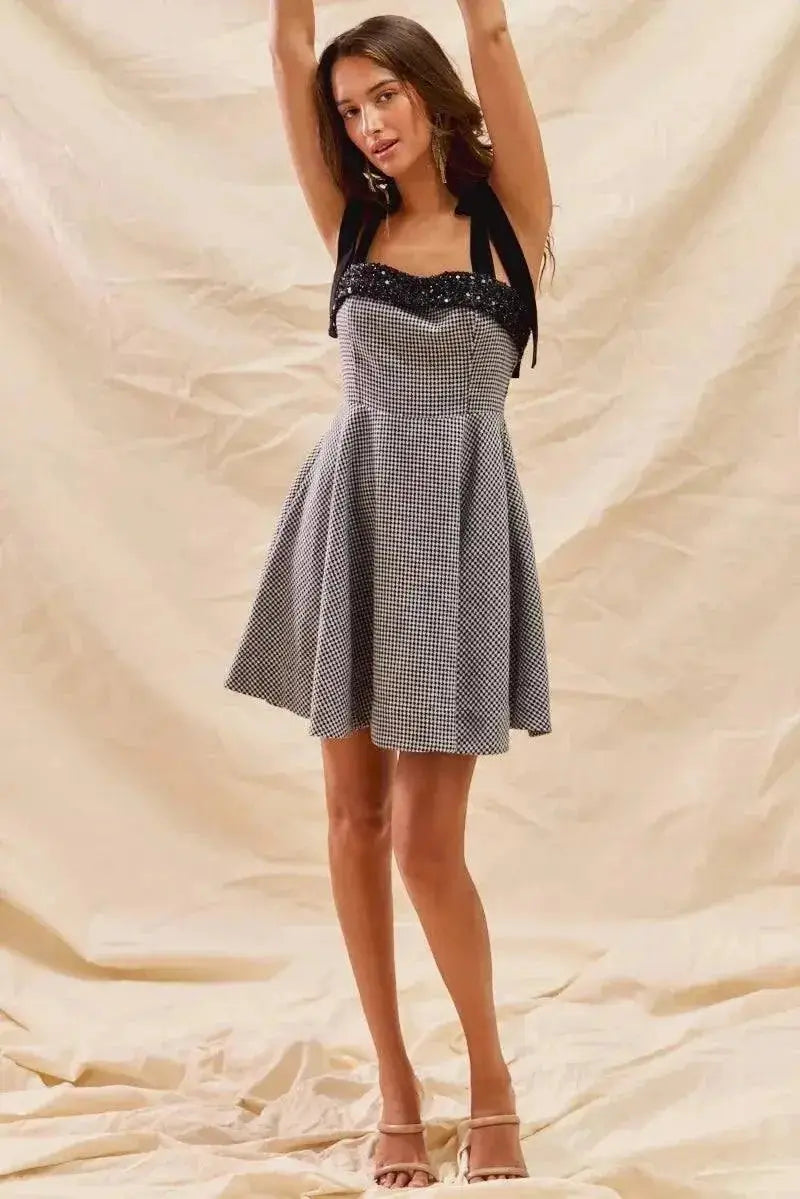 Woman wearing a houndstooth fit flare mini dress with adjustable shoulder ties and sequin detail