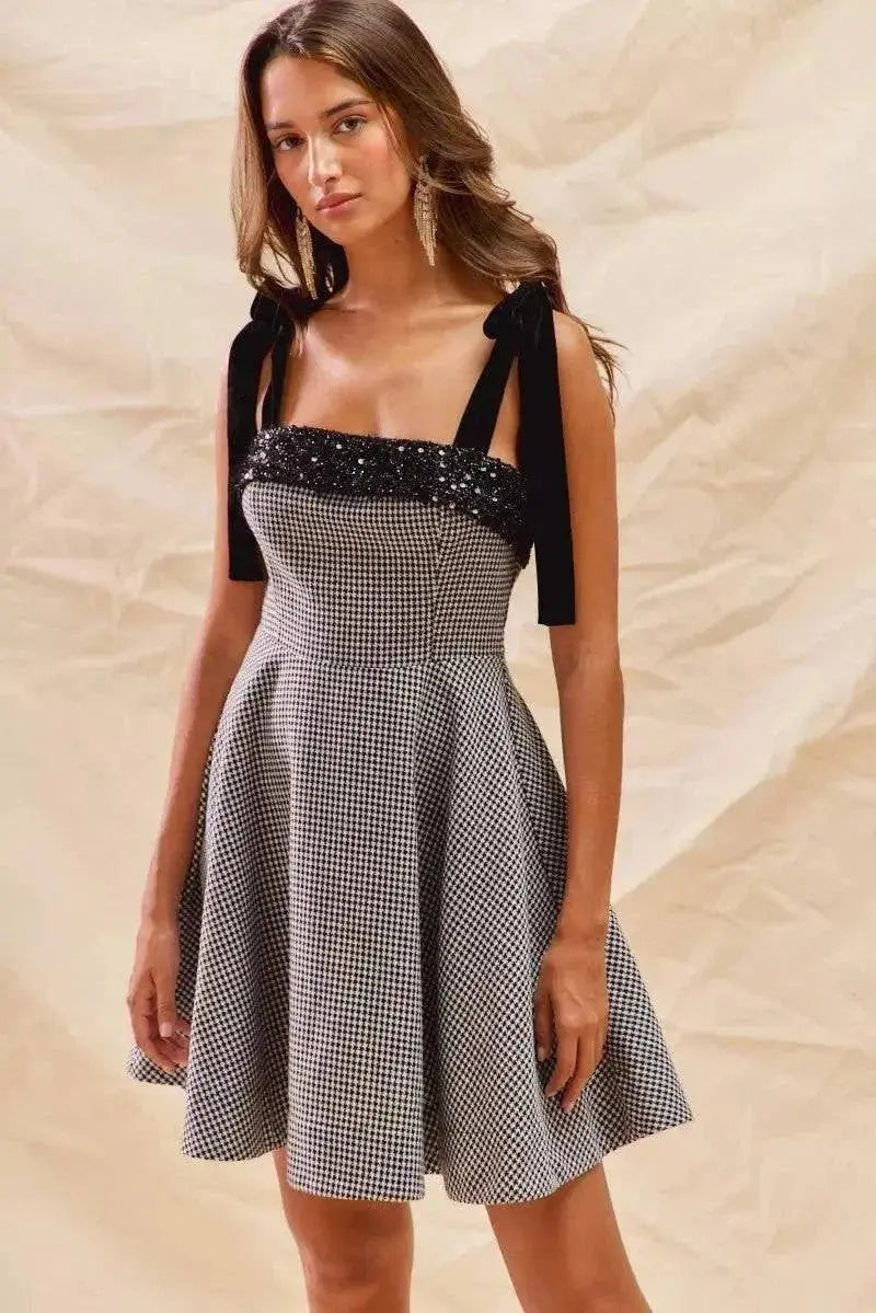 Woman wearing houndstooth fit flare mini dress with black shoulder ties and sequin bodice detail