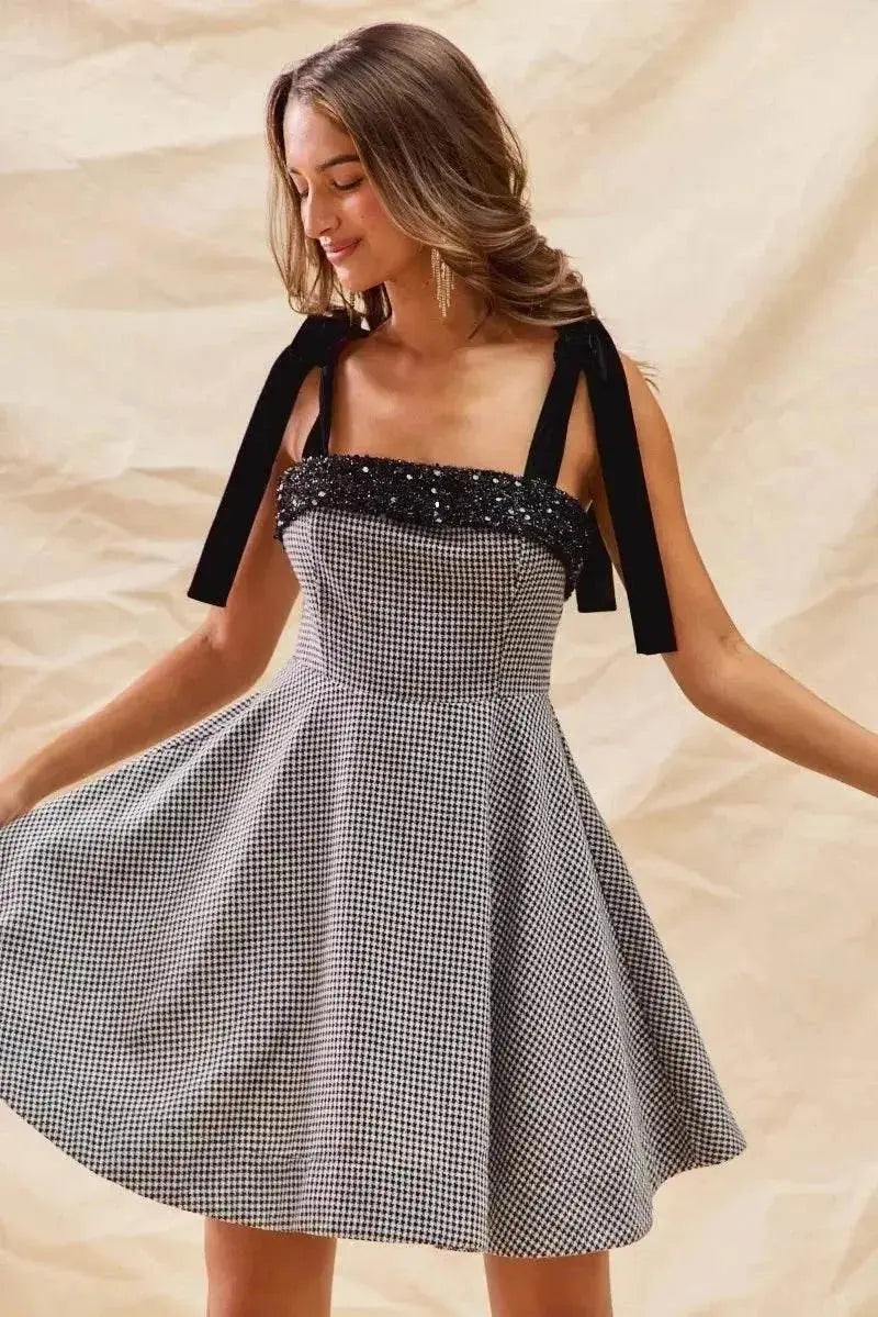 Woman wearing a houndstooth fit flare mini dress with black shoulder ties and sequin detailing